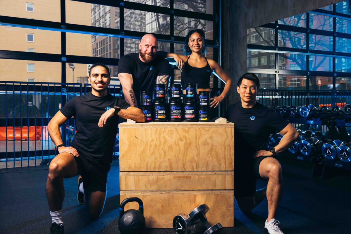 Four fitness professionals posing around Bodybuilding.com supplements