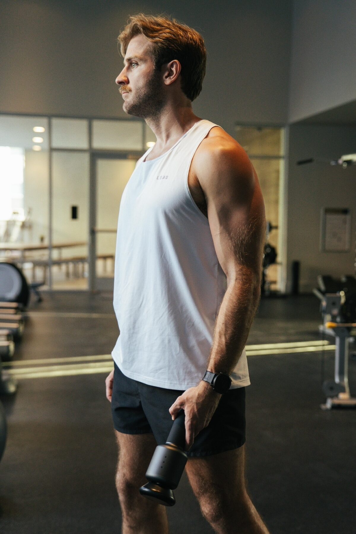 A fit man standing in a gym holding a massage gun